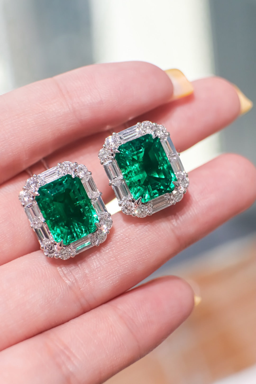 Square emerald stud earrings with diamond halo and baguette accents, shown on fingers